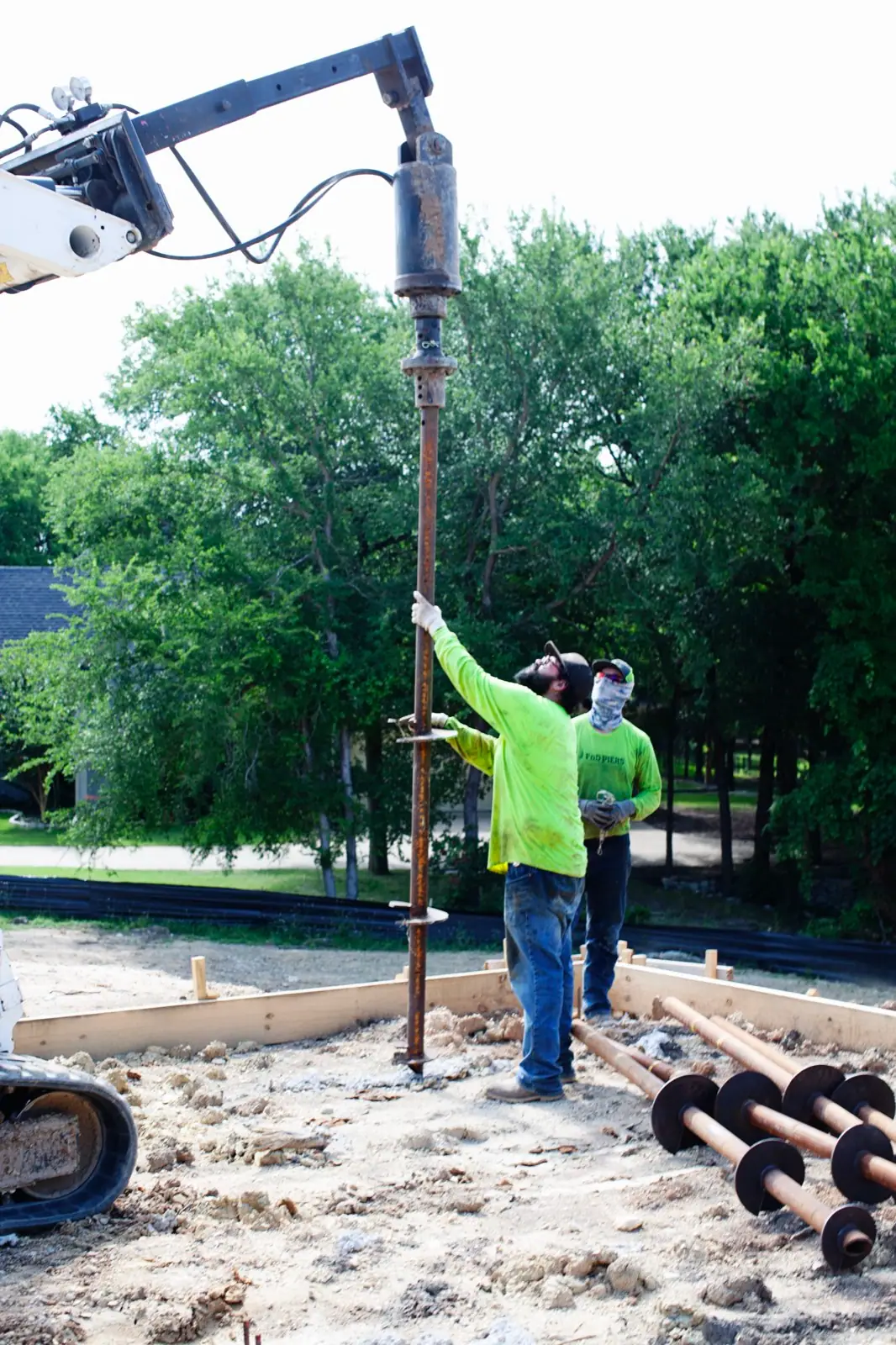 FnD Piers crew guiding a helical pier onto the drive head, with piers staged on the ground