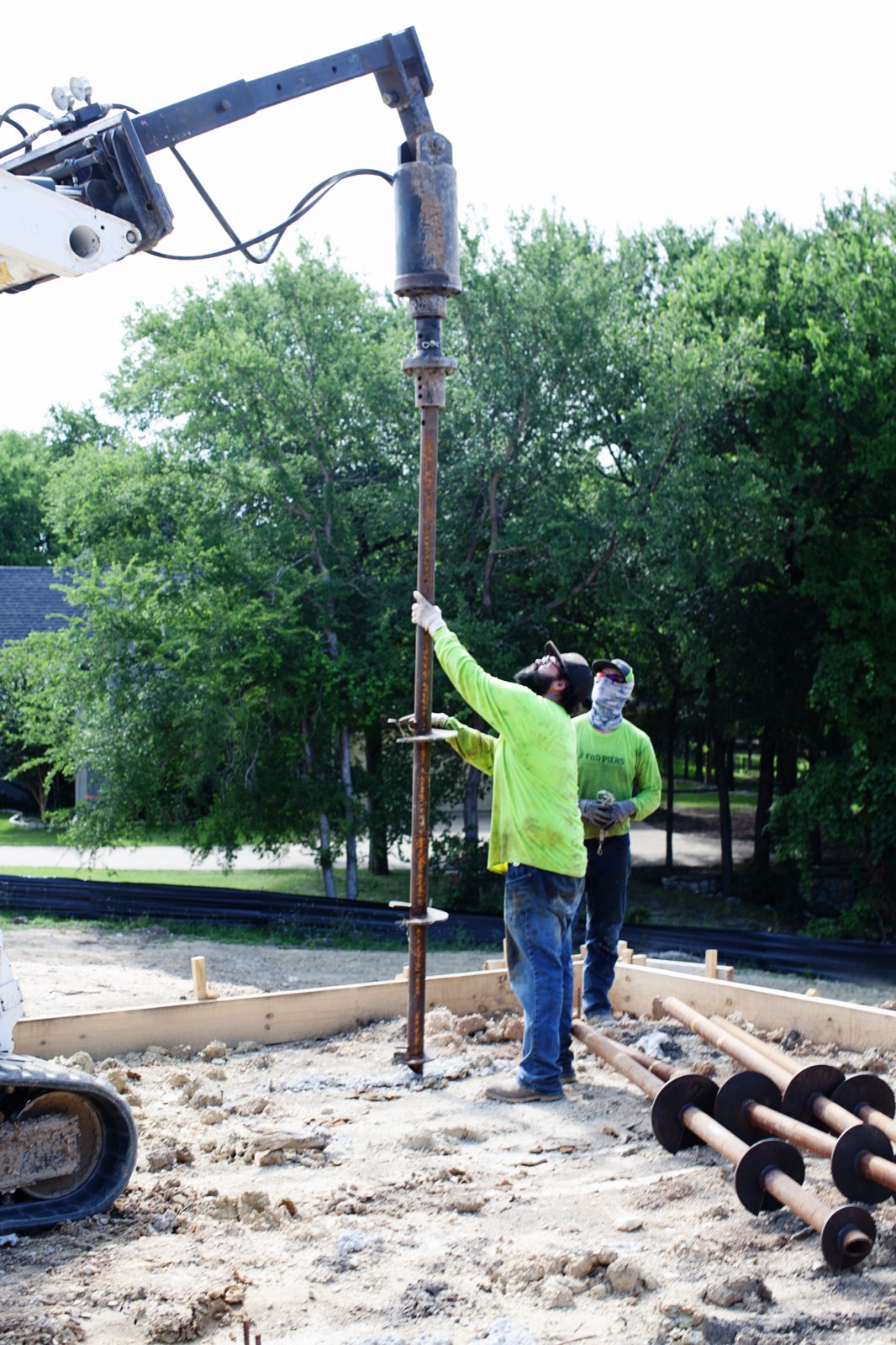 FnD Piers crew guiding a helical pier onto the drive head, with piers staged on the ground